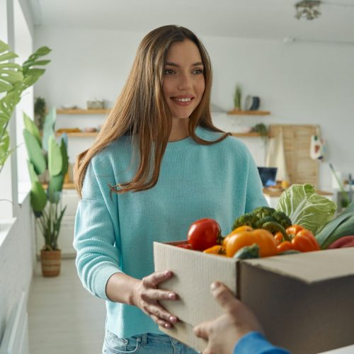 happy young woman accepting box with groceries from delivery man at home